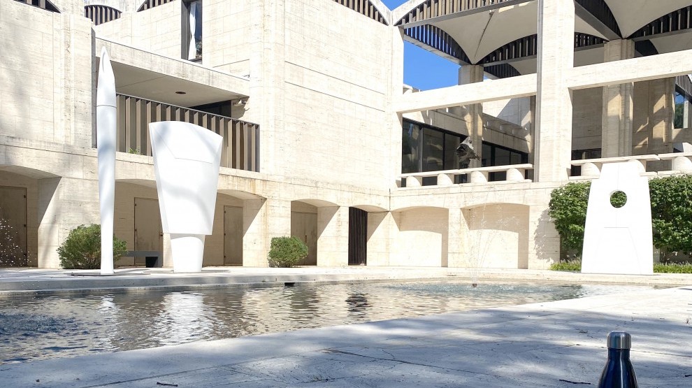 Light beige concrete buildings surround a central water-court flanked by large, white sculptures.